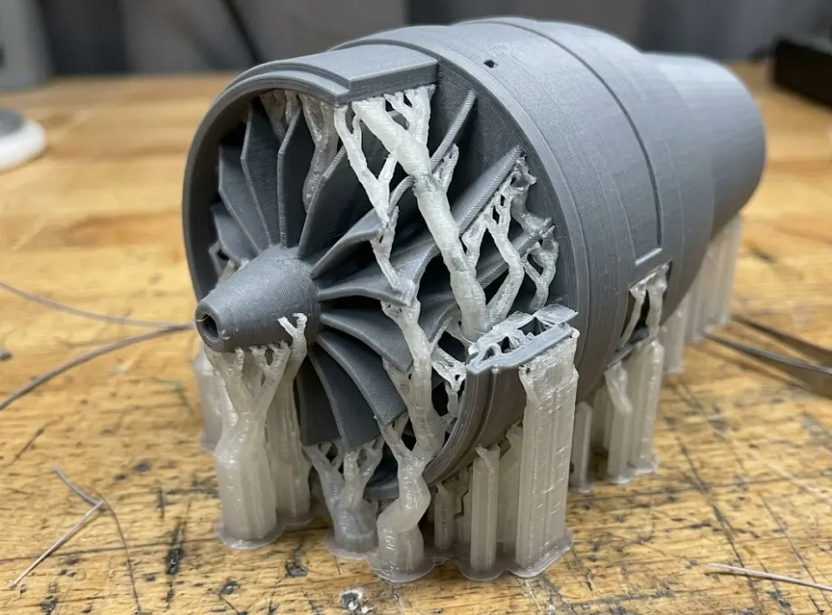 A complex grey 3D printed turbine prototype featuring translucent white soluble support material holding up its internal overhangs.